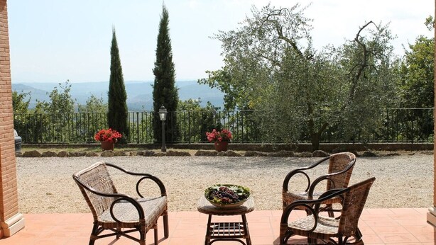 A patio with wicker chairs and a table, overlooking a scenic landscape with trees and a fence.