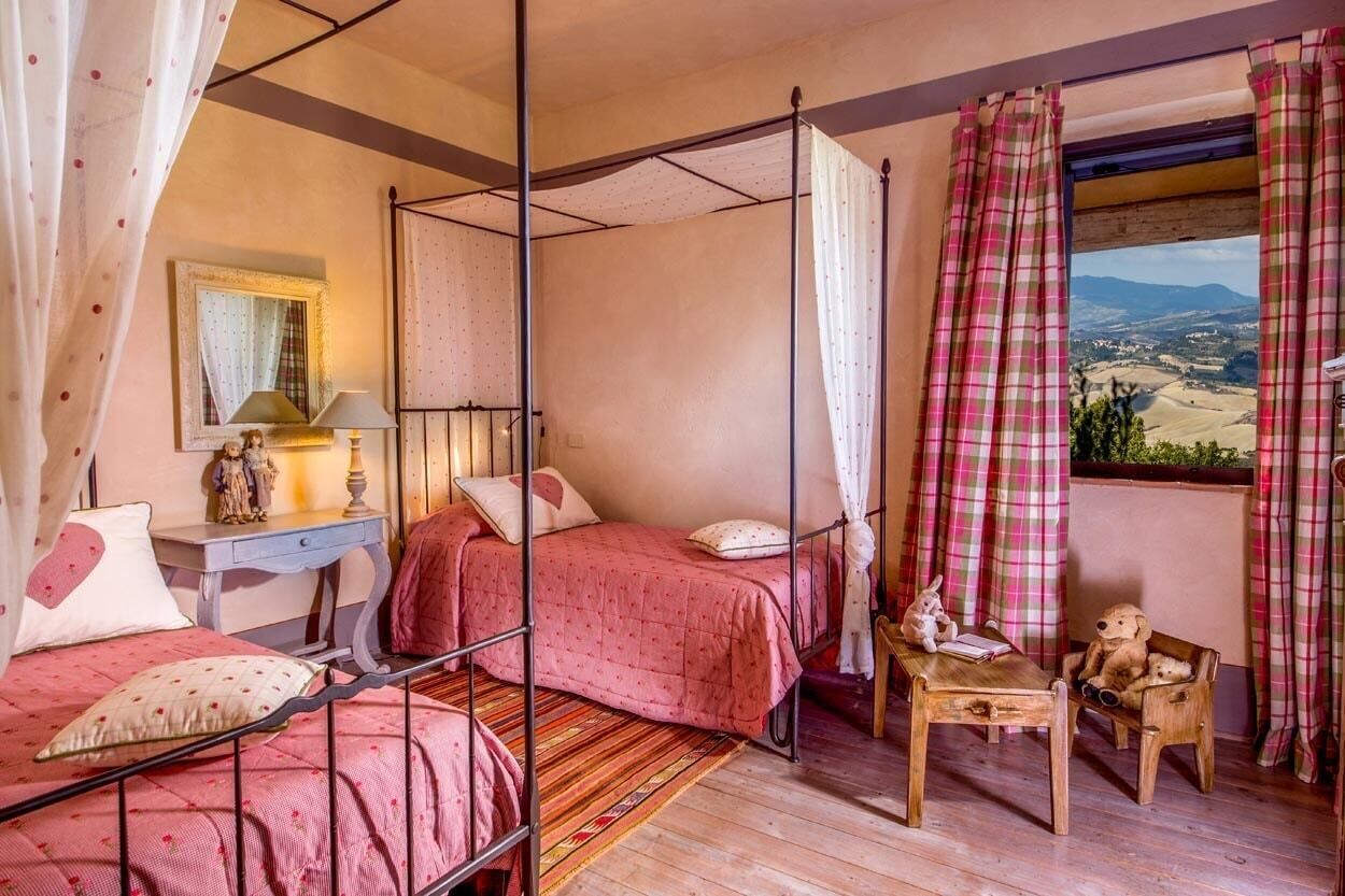 A bedroom with a four-poster bed, a small desk with a lamp, a chair, and a window with a view of the countryside.