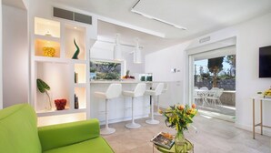 A modern living room with a green sofa, white chairs, a small table with a vase of flowers, and a kitchen area with white bar stools. - Resort Lubrense - perfect for large groups (Sant'Agata Sui Due Golfi)