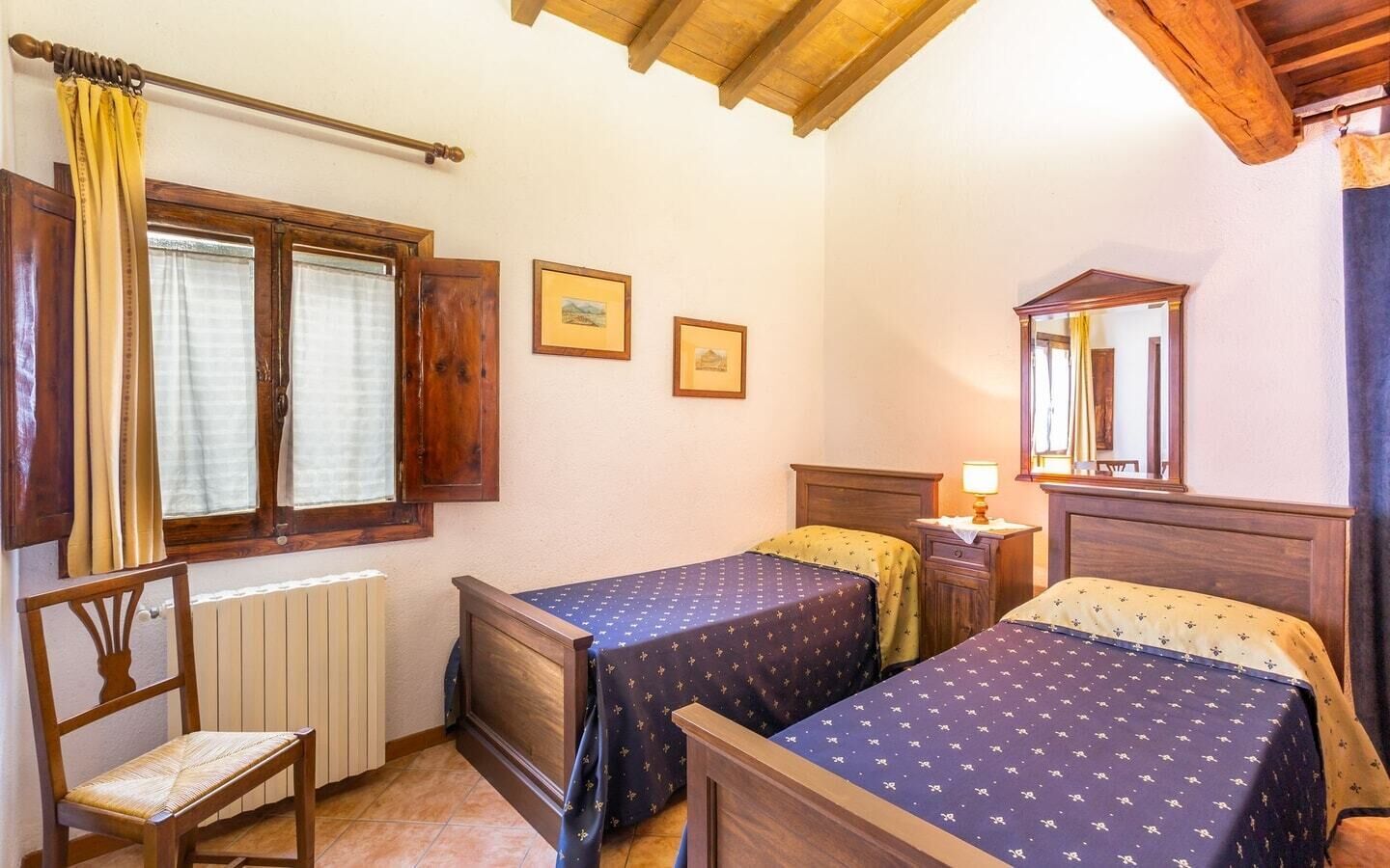 A room with two beds, a wooden chair, a radiator, a window with curtains, and a mirror on the wall.