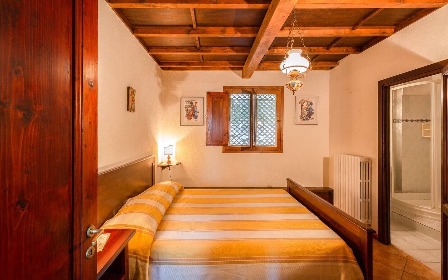 A bedroom with a wooden ceiling, a bed with striped bedding, a nightstand, a wall-mounted lamp, and a window with a lattice design.