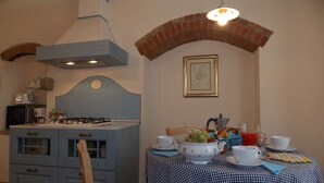 A kitchen with a gas stove, microwave, and blue cabinets. There is a dining table with a checkered tablecloth, a fruit bowl, and a teapot.