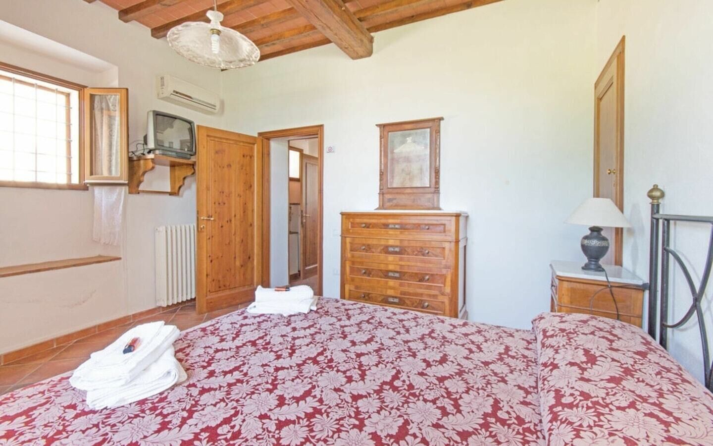 Une chambre avec un plafond en bois, un lit recouvert d’une couvre-lit à motifs floraux, une commode en bois, un miroir et une lampe.