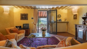 Ein gemütliches Wohnzimmer mit einer Couch, Sesseln, einem Couchtisch und einem Fernseher. Durch eine offene Tür hat man Blick auf einen Garten.