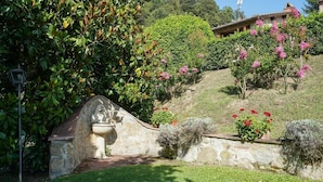 Ein Steinkunstbrunnen, üppiges Grün und blühende Pflanzen in einem Garten.