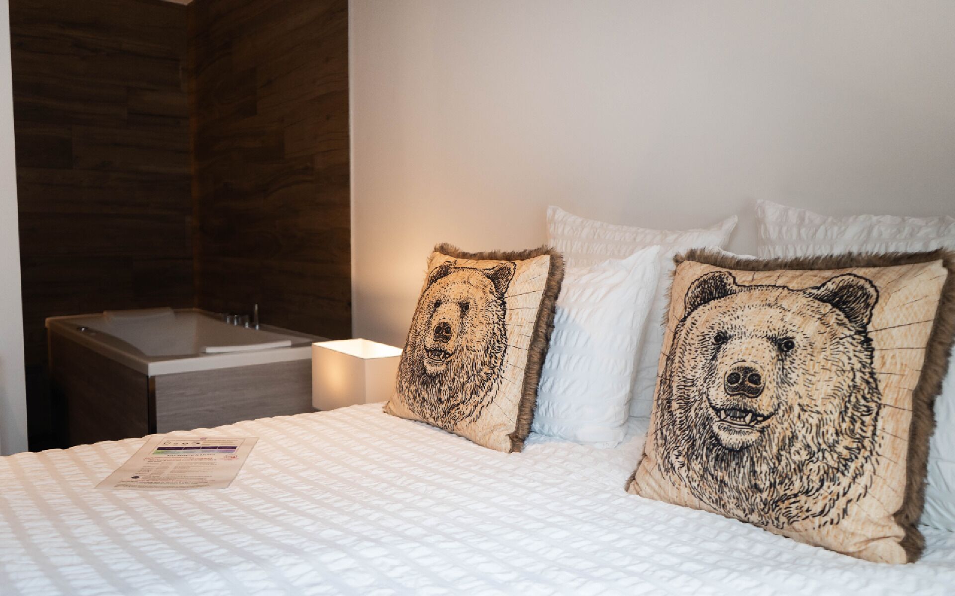 Une chambre d’hôtel avec un lit, deux oreillers ornés de motifs d’ours, une table de chevet et une salle de bain avec un mur en bois et une baignoire.