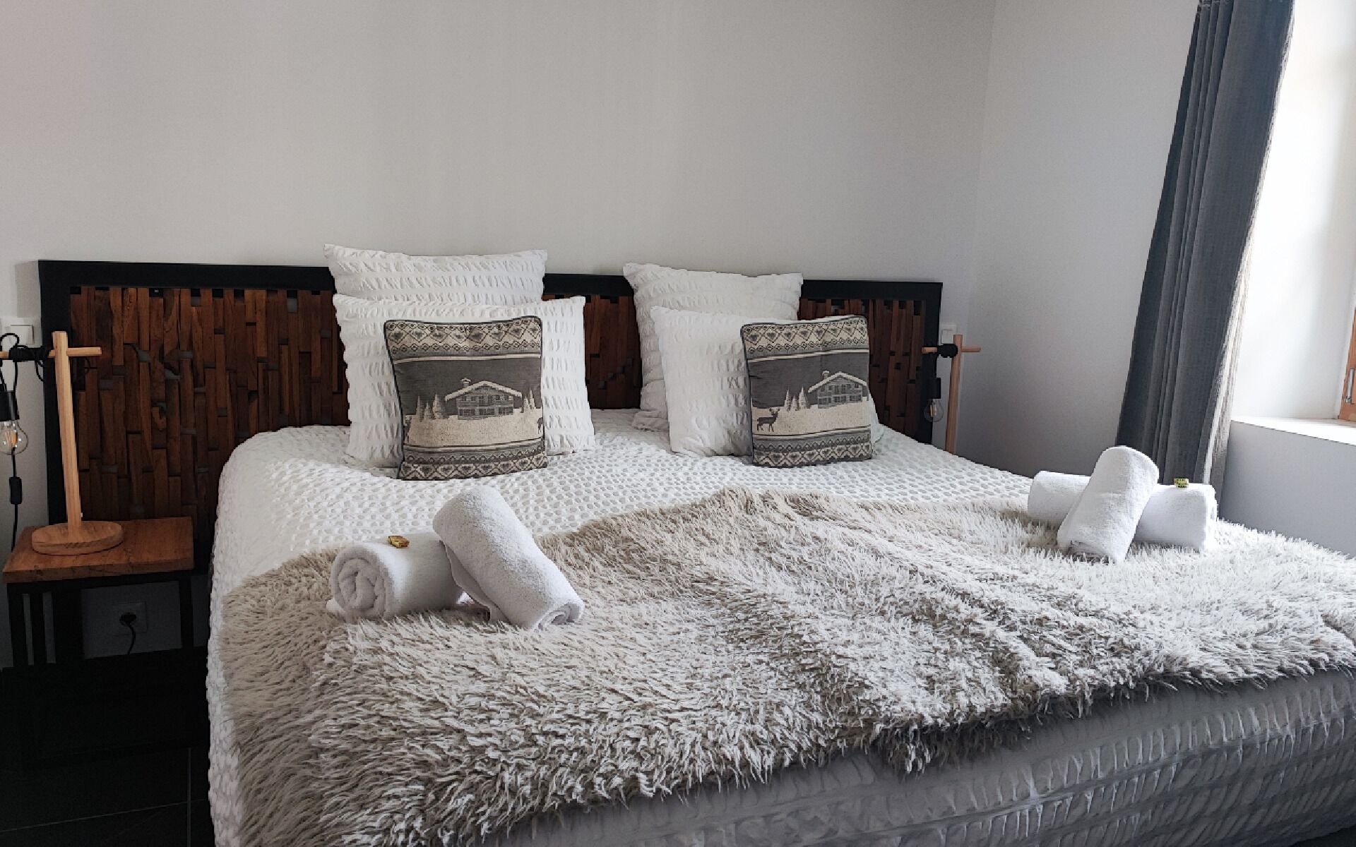 Un lit bien fait, recouvert d’une couverture blanche moelleuse et agrémenté de deux coussins décoratifs, avec une tête de lit en bois et une table de chevet sur laquelle se trouve une lampe.