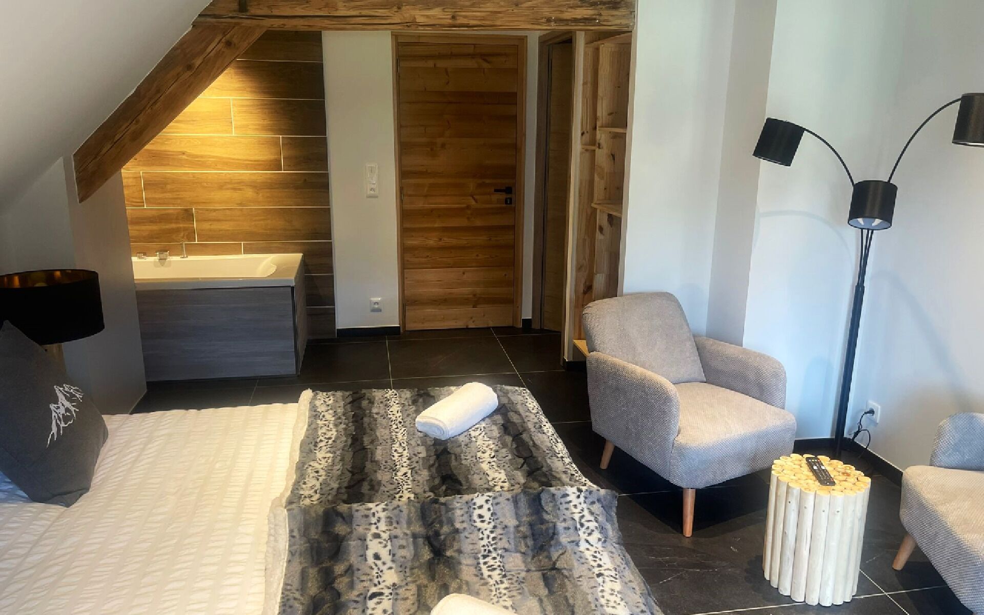 Un dormitorio moderno con techo inclinado, bañera integrada, un sillón gris, una lámpara de pie y una mesa de centro original.