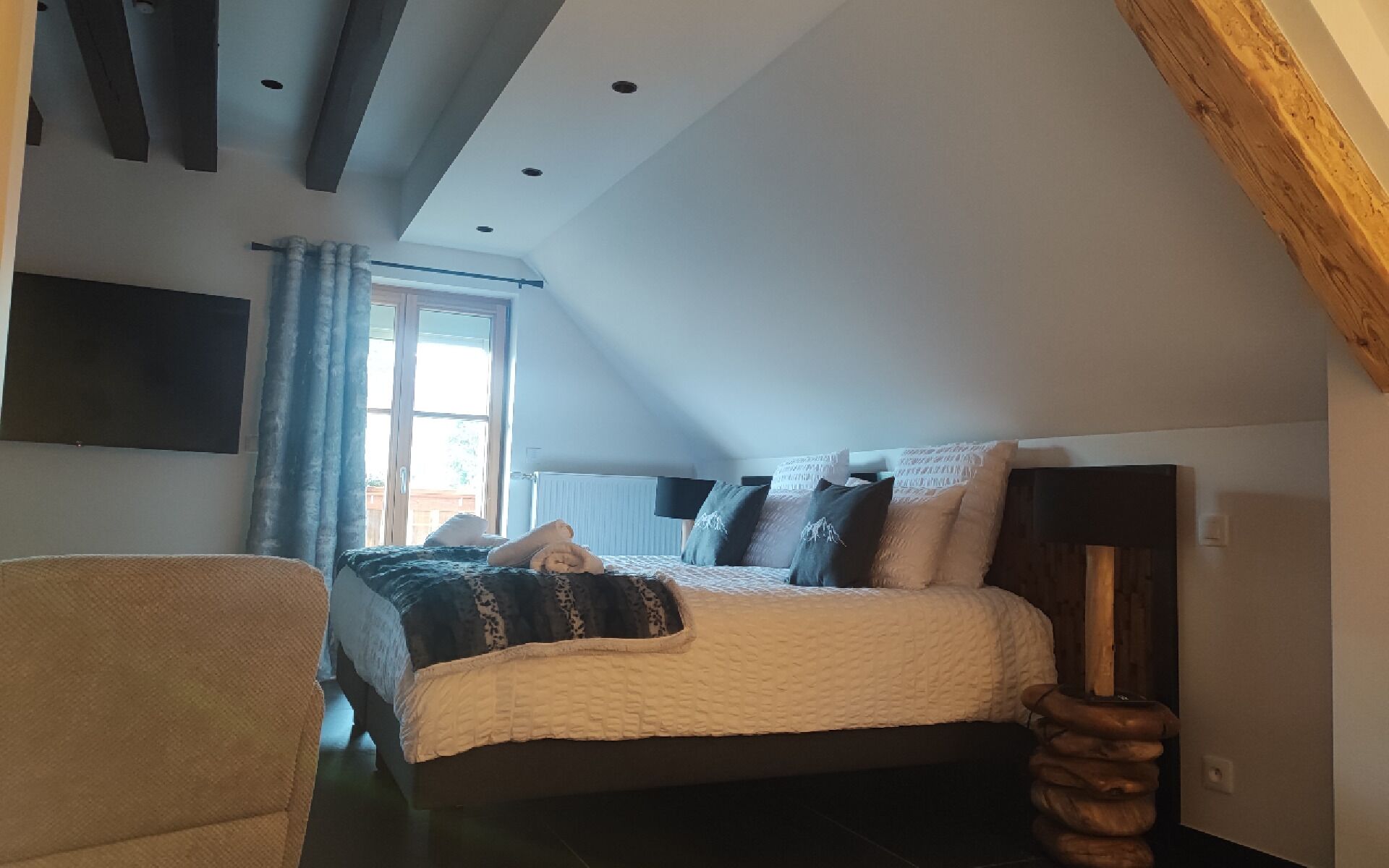 Ein Schlafzimmer mit Bett, Sessel, Holz-Kopfteil, Fenster und einem angebrachten Fernseher.
