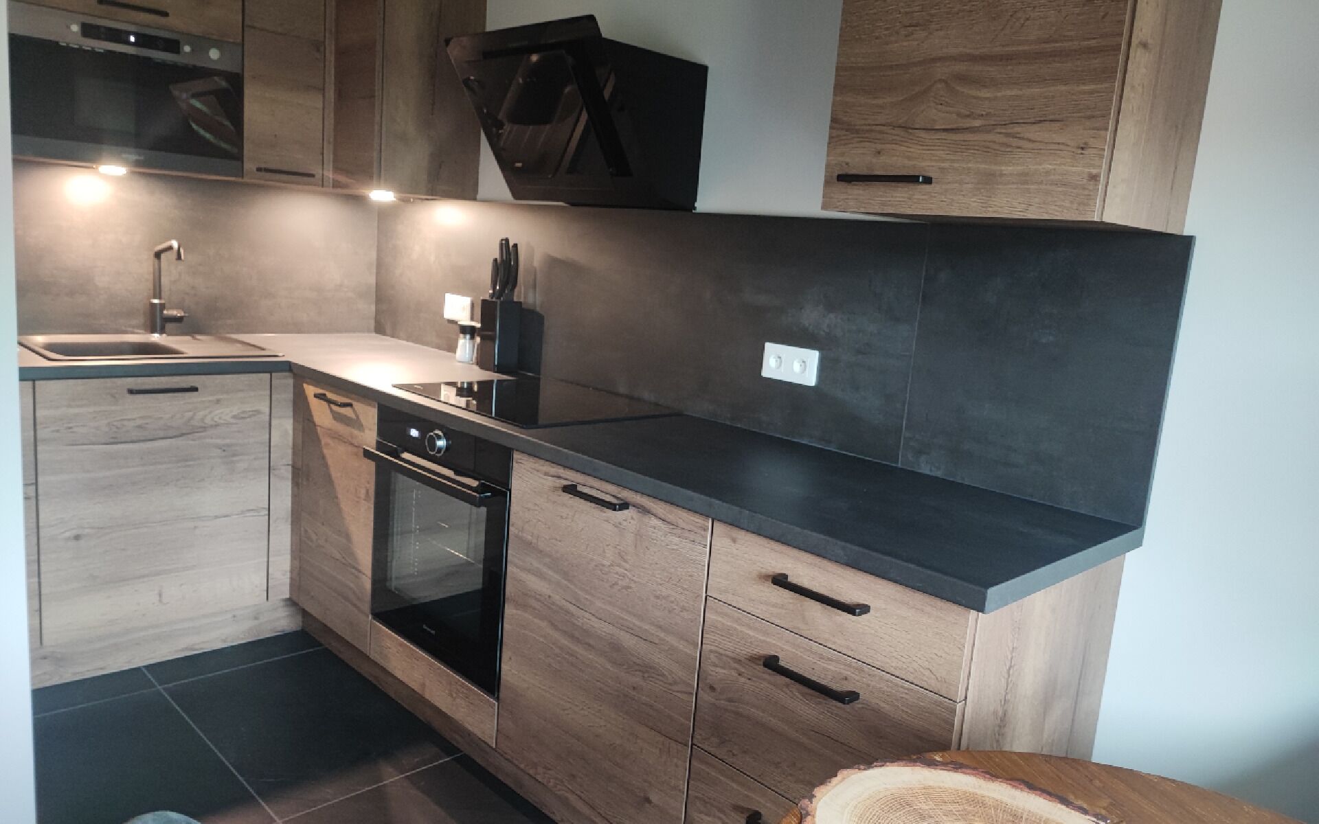 Una cocina moderna con armarios de madera, encimera negra, horno integrado y un lavamanos.
