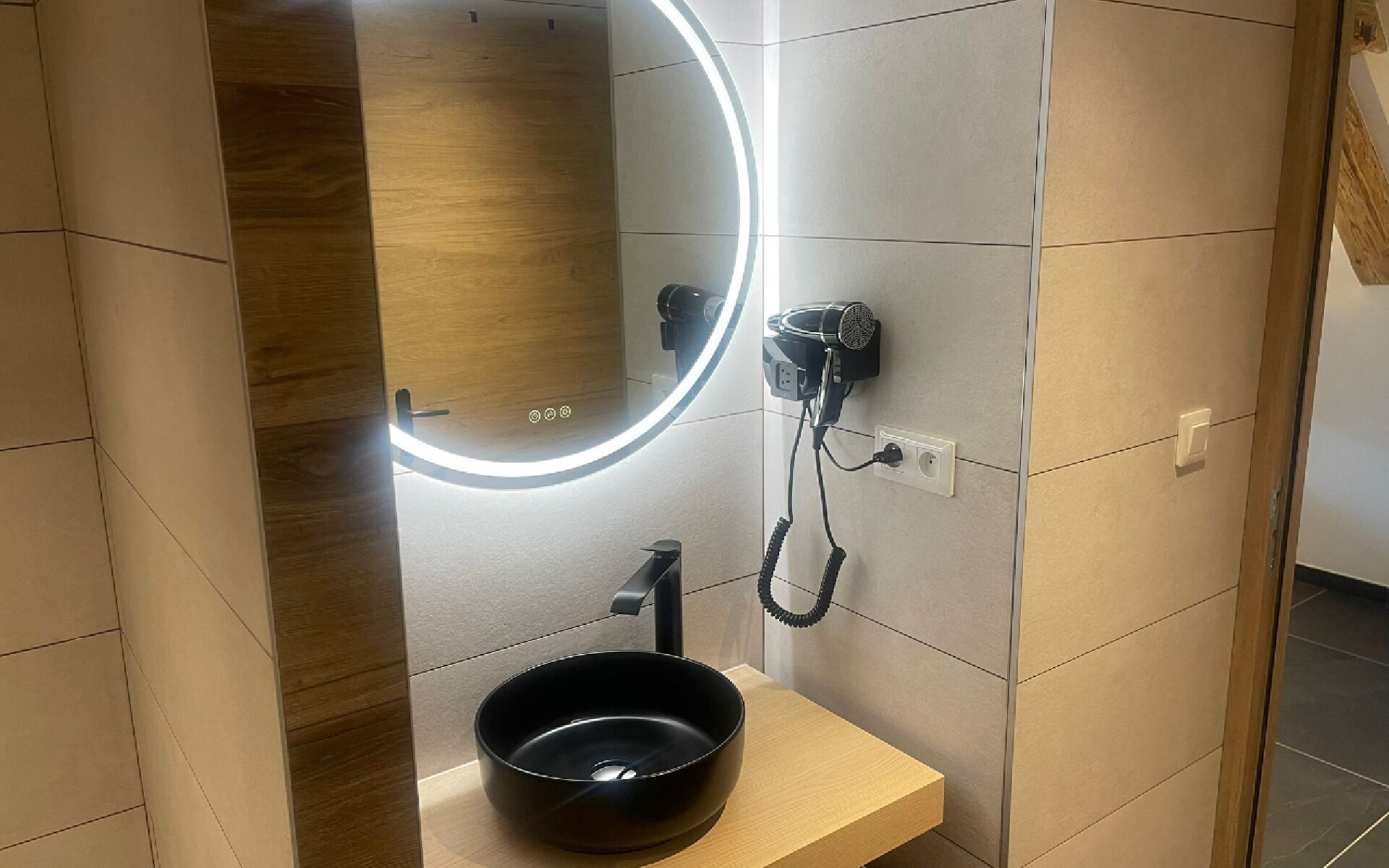 A bathroom with a modern design, featuring a wooden vanity, a black sink, a hairdryer, and a mirror with a lighted frame.