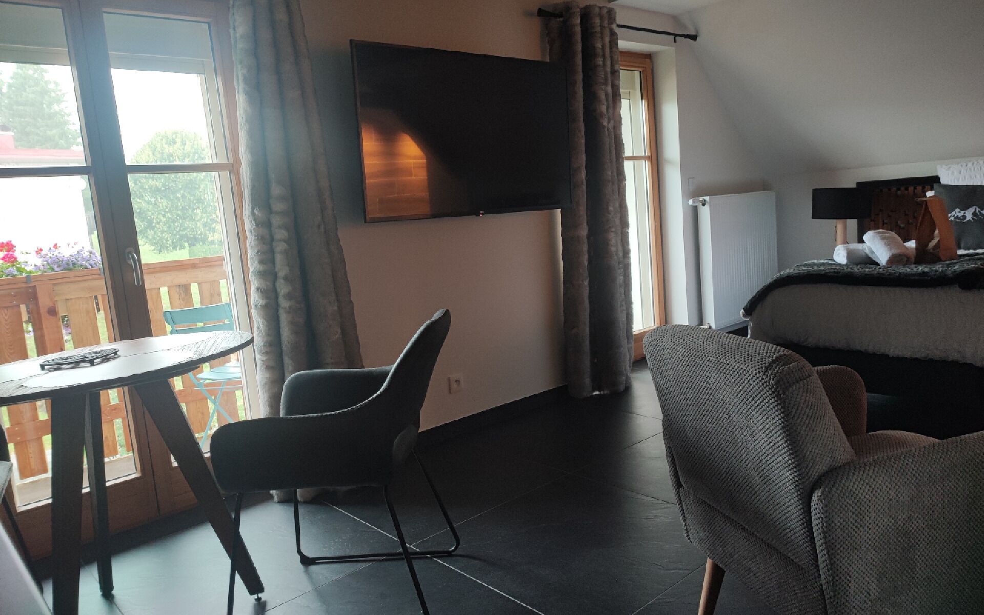 Une chambre d’hôtel avec un lit, un téléviseur fixé au mur, une petite table avec une chaise, et une vue sur l’extérieur grâce à de grandes fenêtres.
