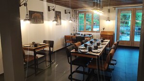 Un espace repas comprenant une longue table, plusieurs chaises et un banc. Des lampes sont fixées au mur, et une fenêtre offre une vue sur la verdure à l’extérieur.