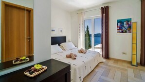 A hotel room with a bed, a desk with a basket of fruit, a large window with a view of the sea, and a painting on the wall.