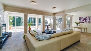 A spacious living room with a beige sofa, a coffee table, and a TV. There are large windows and a balcony with potted plants.