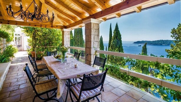 A covered outdoor dining area with a table set for four, overlooking a scenic view of the sea and hills.