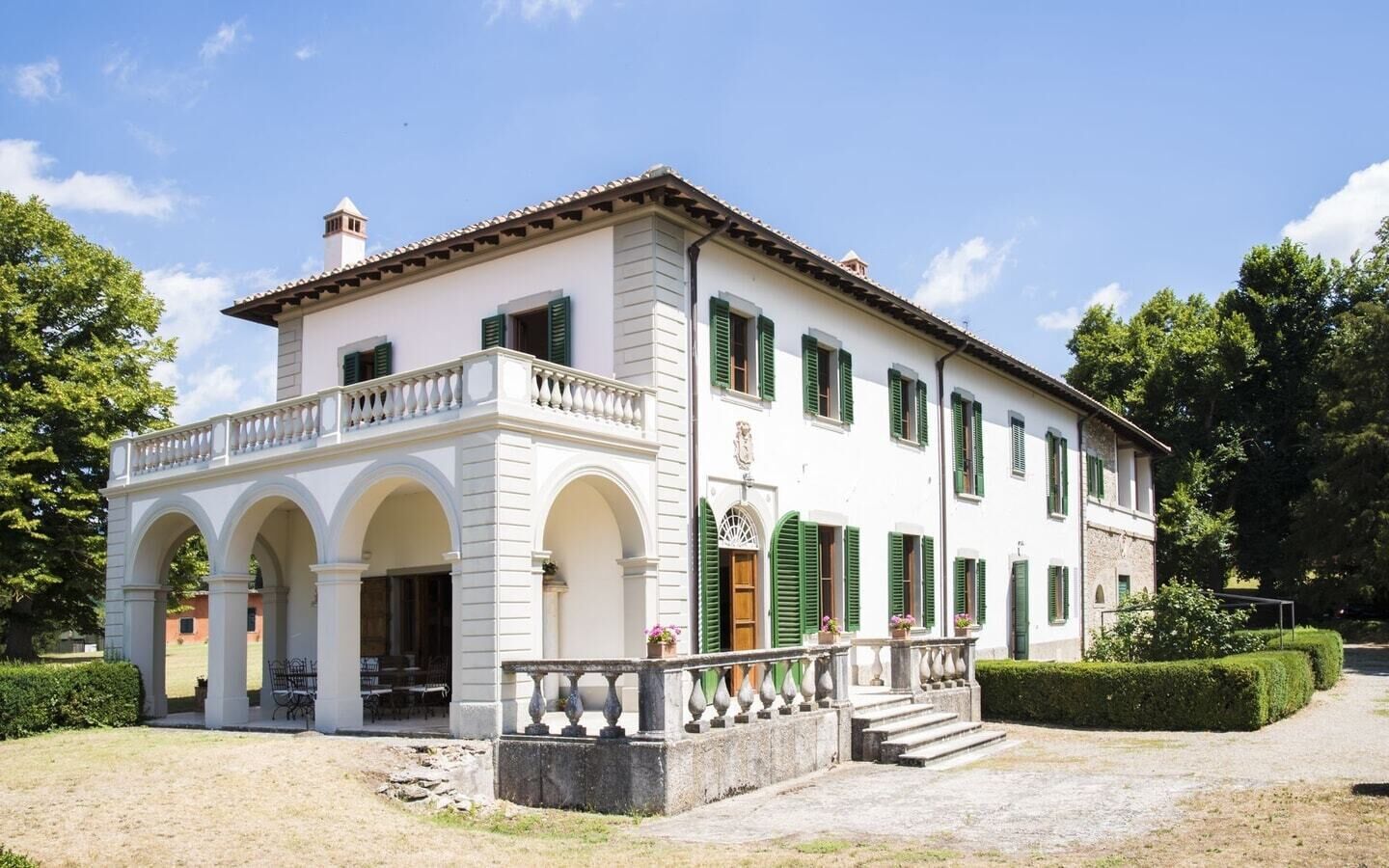 A large, white building with green shutters, a balcony, and a central entrance with steps.