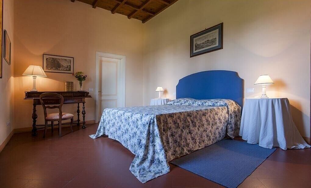 Een slaapkamer met een bed, een bureau, twee stoelen, een deur en een lijst met een afbeelding aan de muur.
