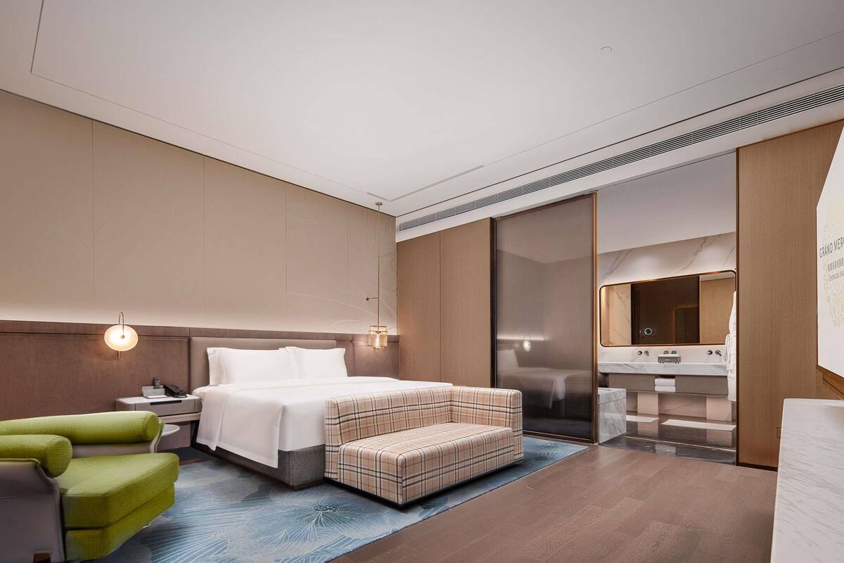 A modern hotel room with a large bed, a green armchair, a small ottoman, a vanity with a mirror, and a wall-mounted light fixture.