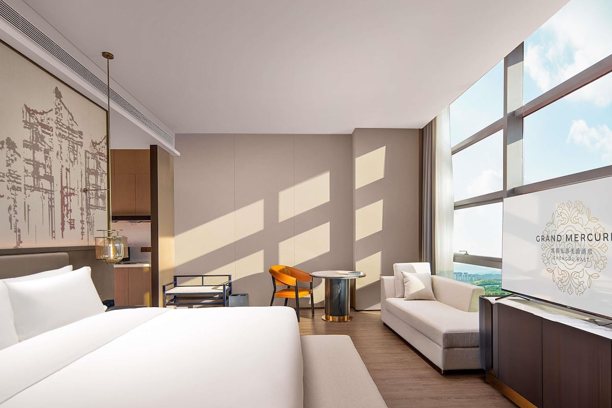 A hotel room with a bed, a sofa, a small table, and a chair. There is a large window with a view of the city.