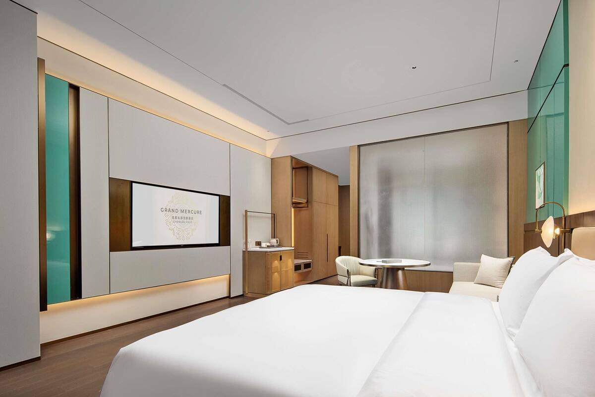 A modern hotel room with a large bed, a desk, a chair, and a television displaying 'GRAND MERCURY CHENGDUO EAST'.