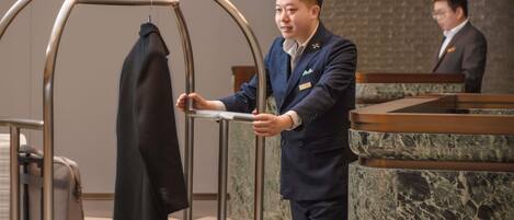 En man i uniform hänger upp en jacka på en metallklädhängare i en entréhall med ett rutmönstrat golv och väggar av marmor.