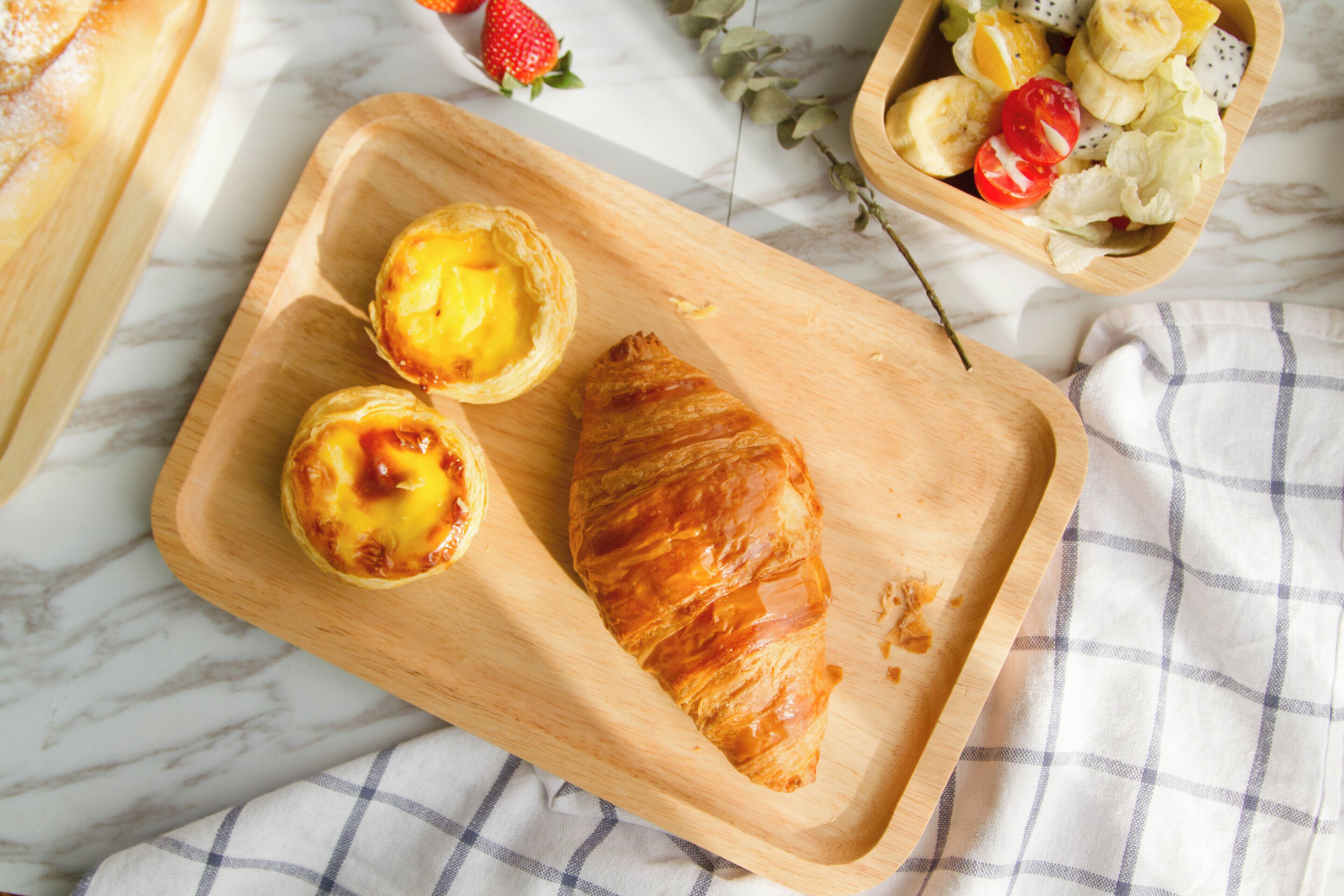 A wooden serving tray with a croissant and a Portuguese egg tart on a marble surface.