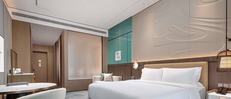 A modern hotel room with a large bed, a desk, a chair, and a bedside table with a lamp.