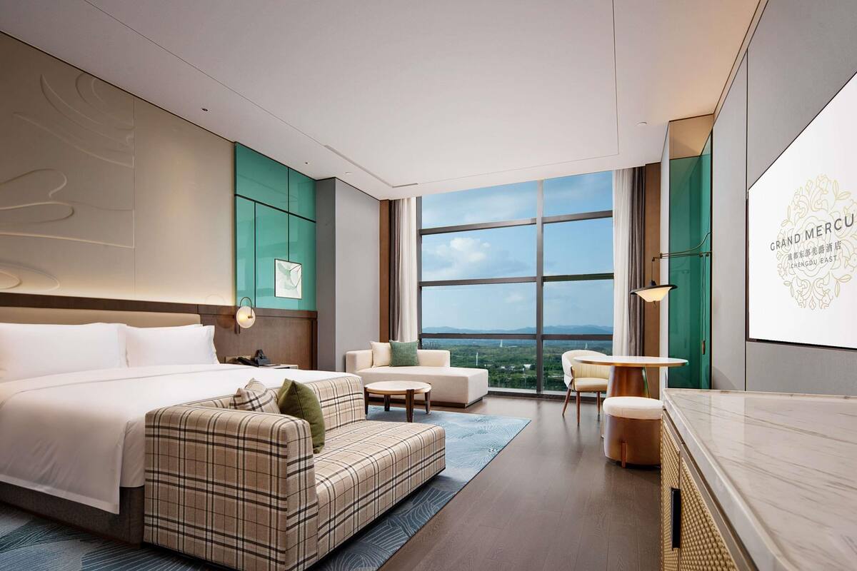 A modern hotel room with a large bed, a sofa, a small table, and a view of the cityscape.