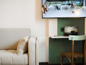 A modern living room with a sofa, a wooden chair, a flat-screen TV mounted on the wall, and a laptop on a stand. - Greet Hotel Brest Aéroport (Guipavas)