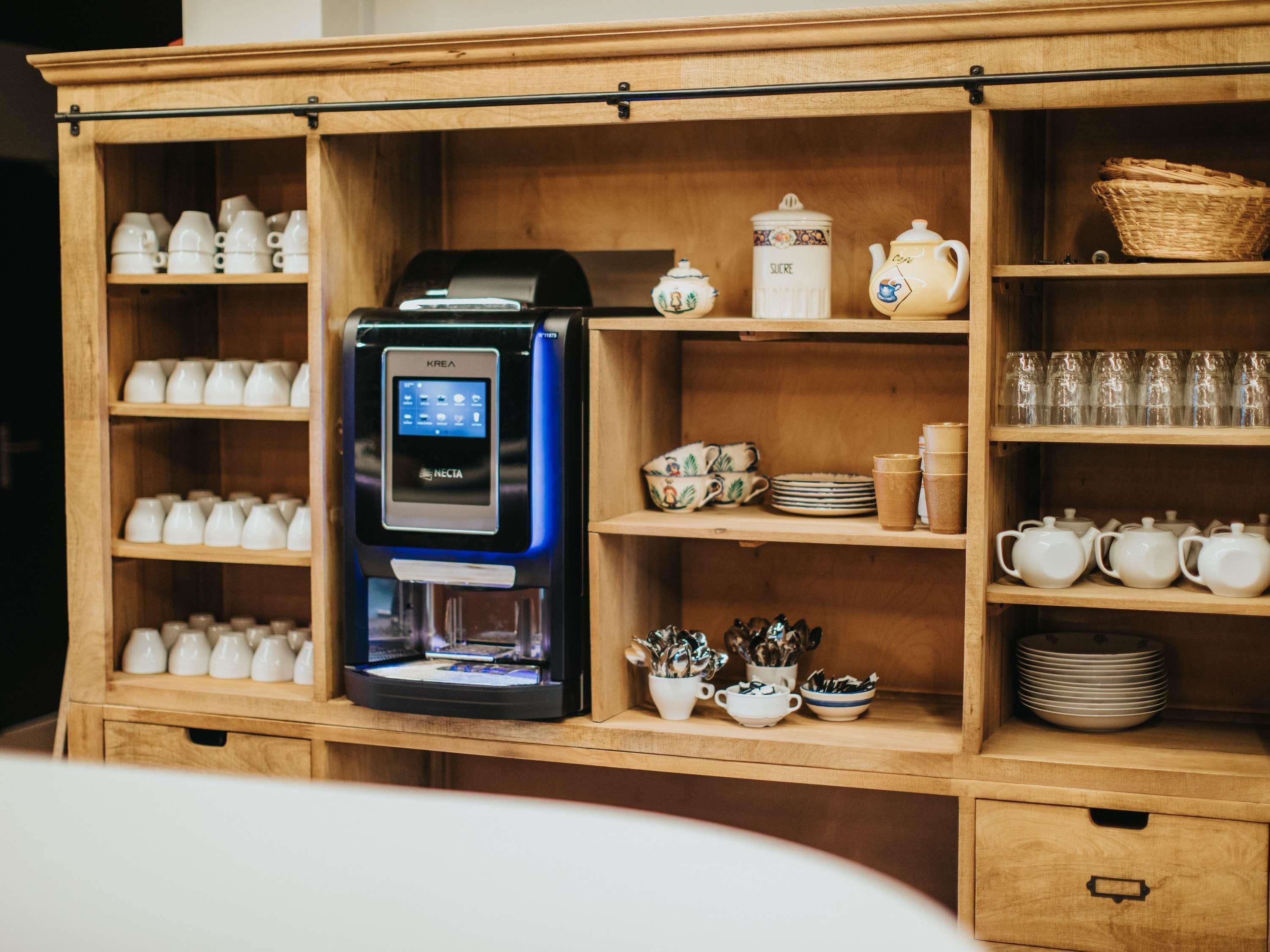 Een koffieapparaat met een digitaal scherm, omringd door planken met verschillende kopjes, borden en theepotten.