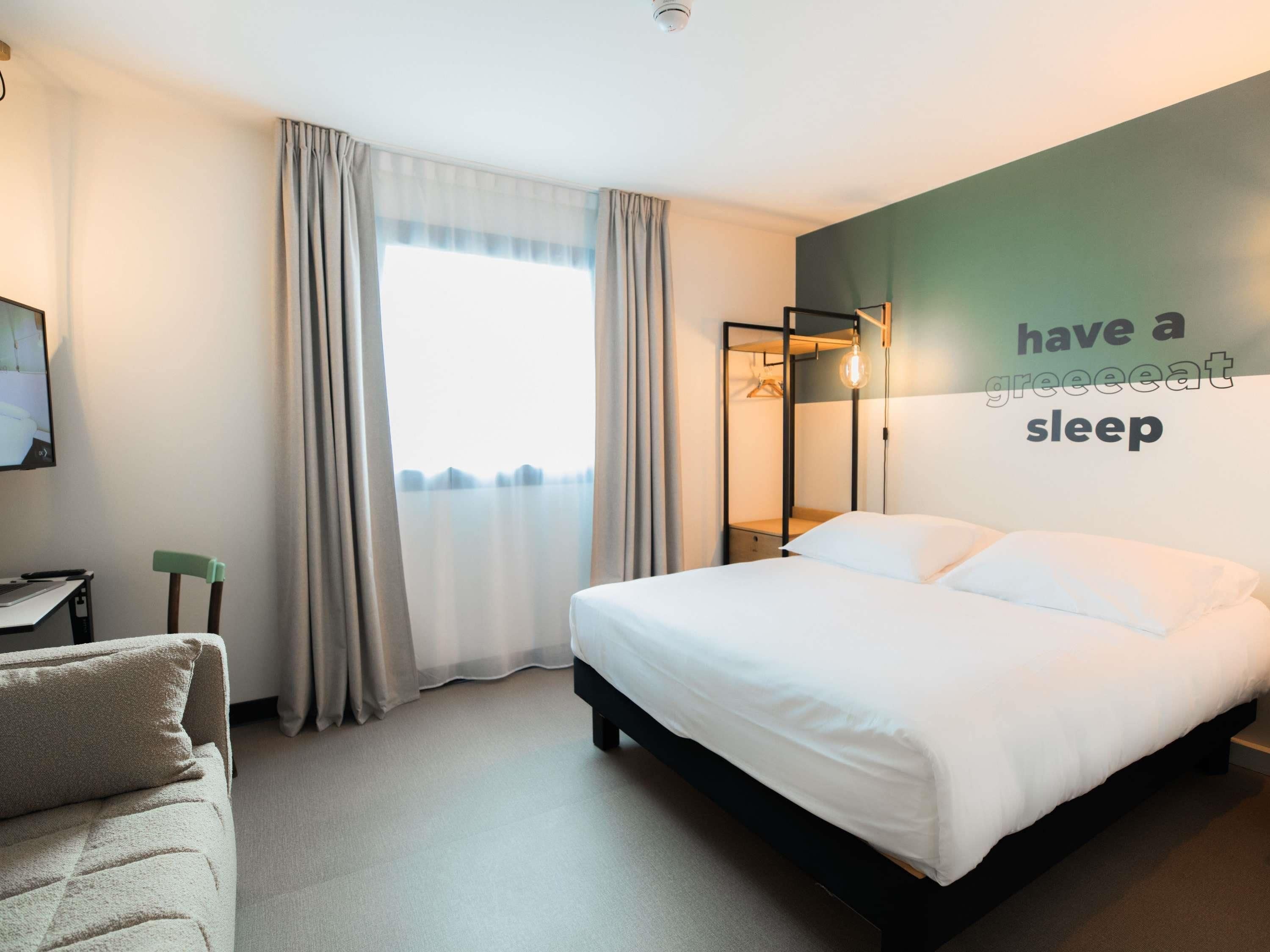 A hotel room with a bed, a desk, a chair, a wardrobe, and a wall with a motivational quote.