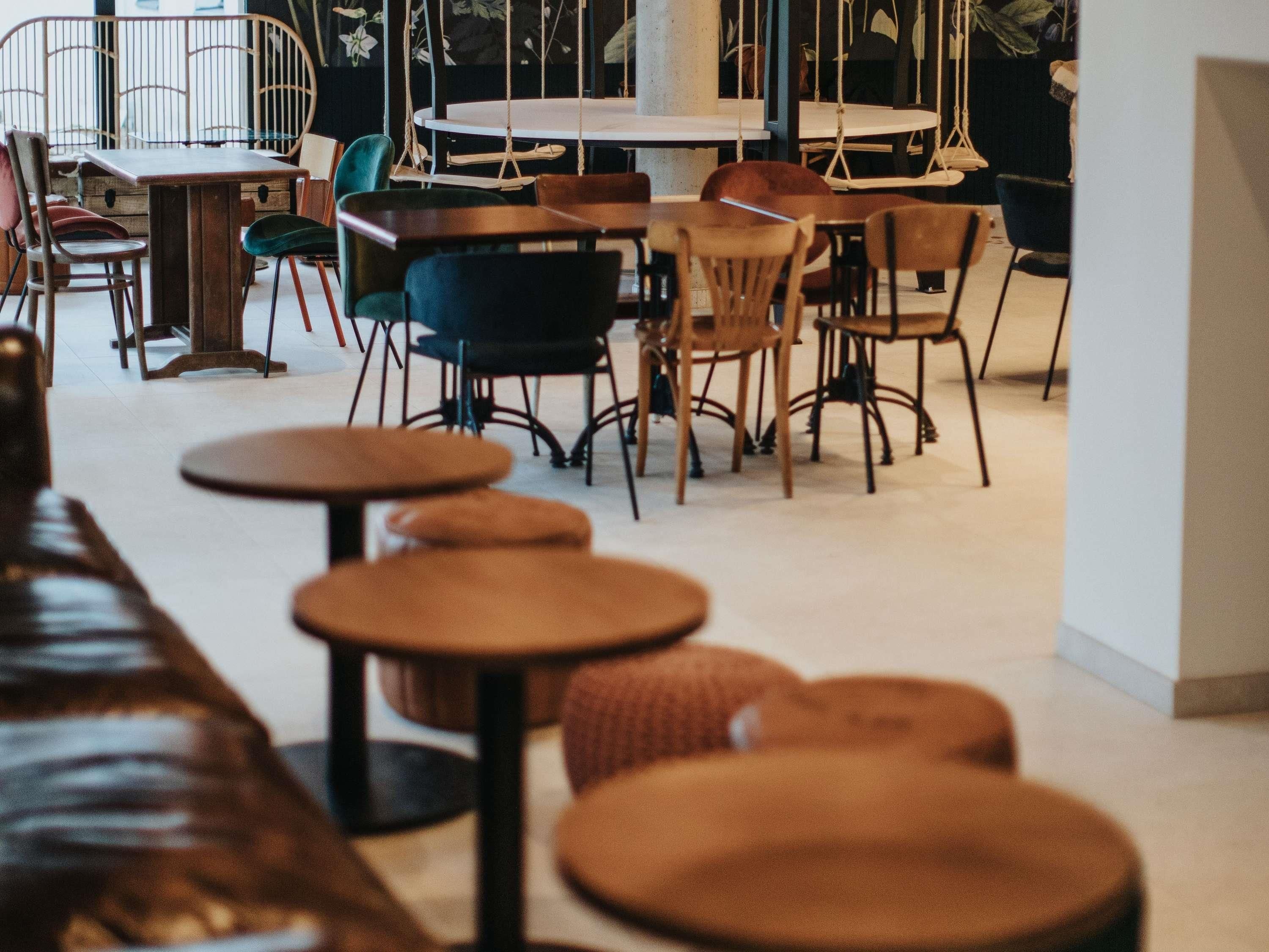 Interior de una cafetería con mesas y sillas redondas, un banco de madera y plantas colgantes.