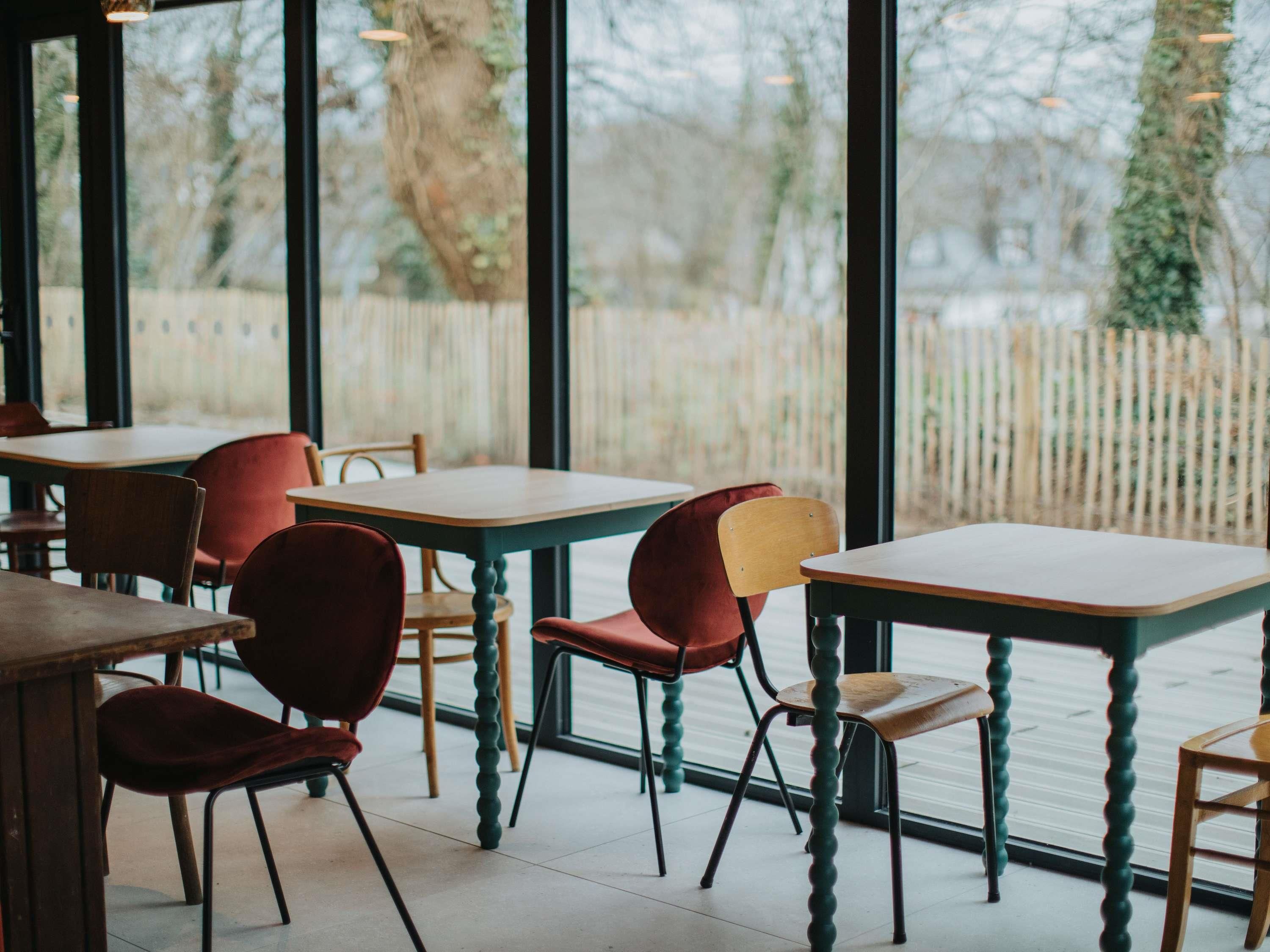 Ein Restaurantinnenraum mit Tischen, Stühlen, großen Fenstern und Blick auf einen Holenzaun und Bäume.
