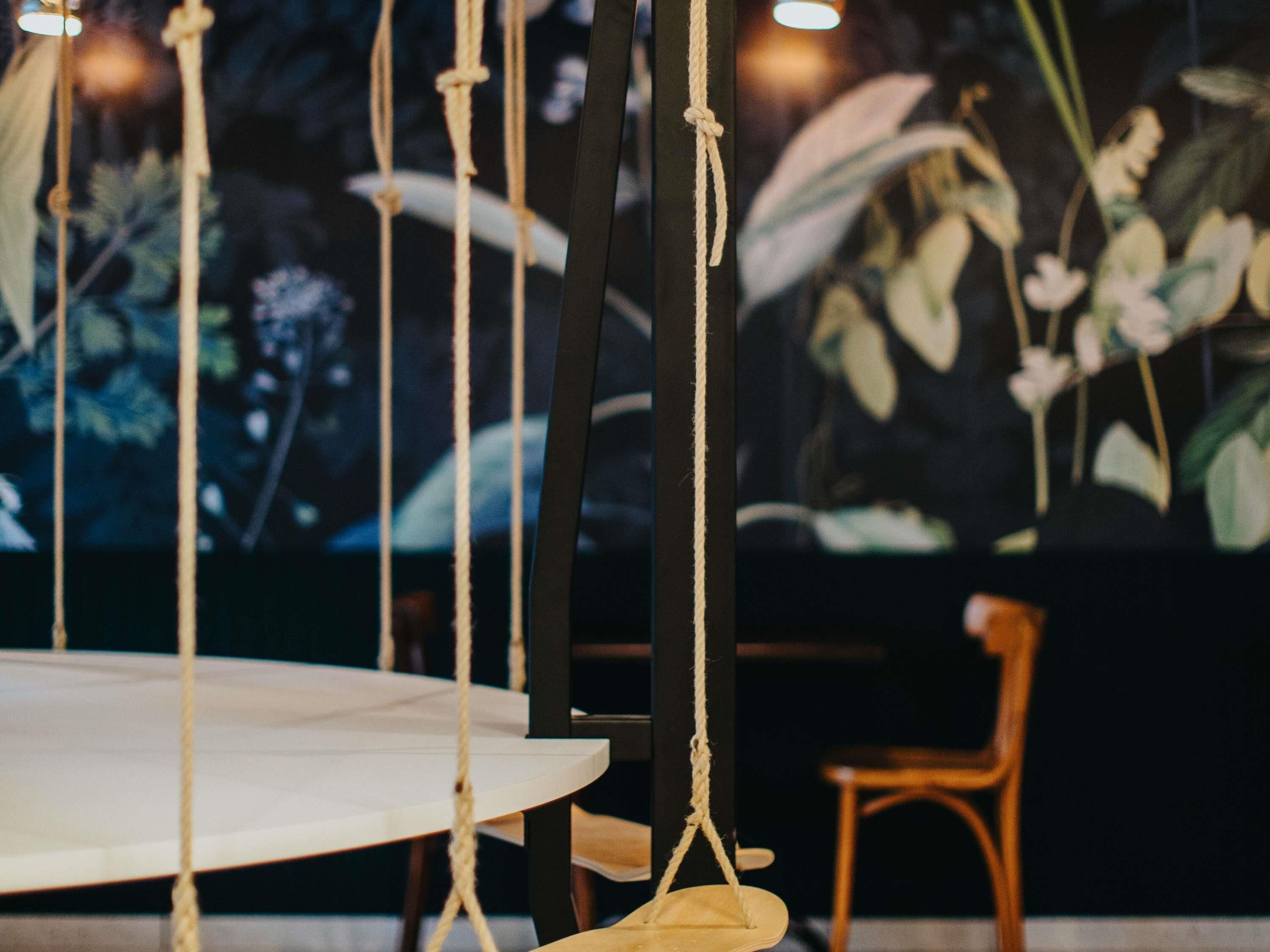 Uma mesa redonda com uma cadeira suspensa em corda, uma cadeira de madeira e uma parede com um mural floral.