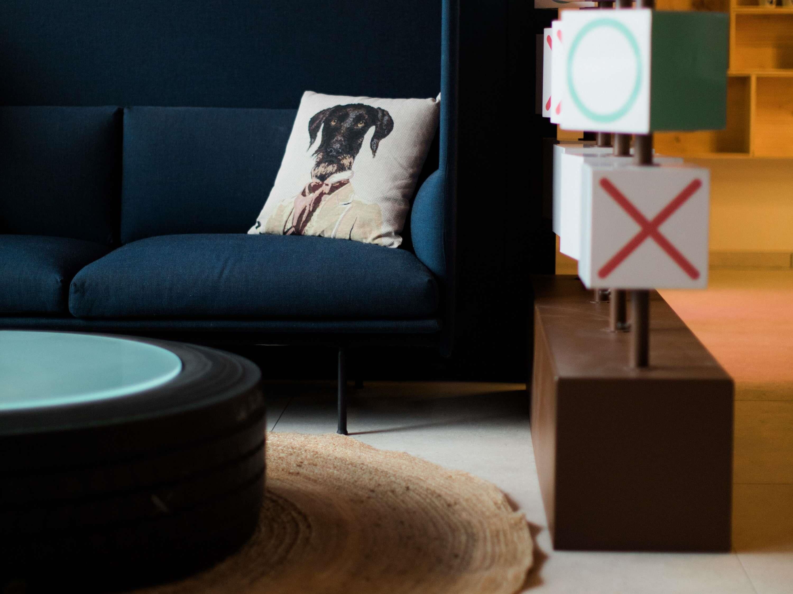 Un sofá azul oscuro con un cojín decorativo, una mesa redonda y un bloque de madera con una 'X' roja.