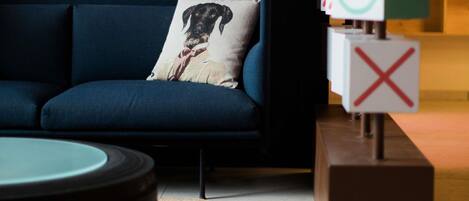 Un canapé bleu foncé agrémenté d’un coussin décoratif, une table ronde et un bloc de bois sur lequel est gravé un « X » rouge.