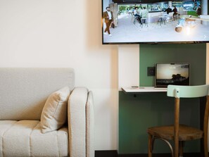 A modern living room with a sofa, a wooden chair, a flat-screen TV mounted on the wall, and a laptop on a stand. - Greet Hotel Brest Aéroport (Guipavas)