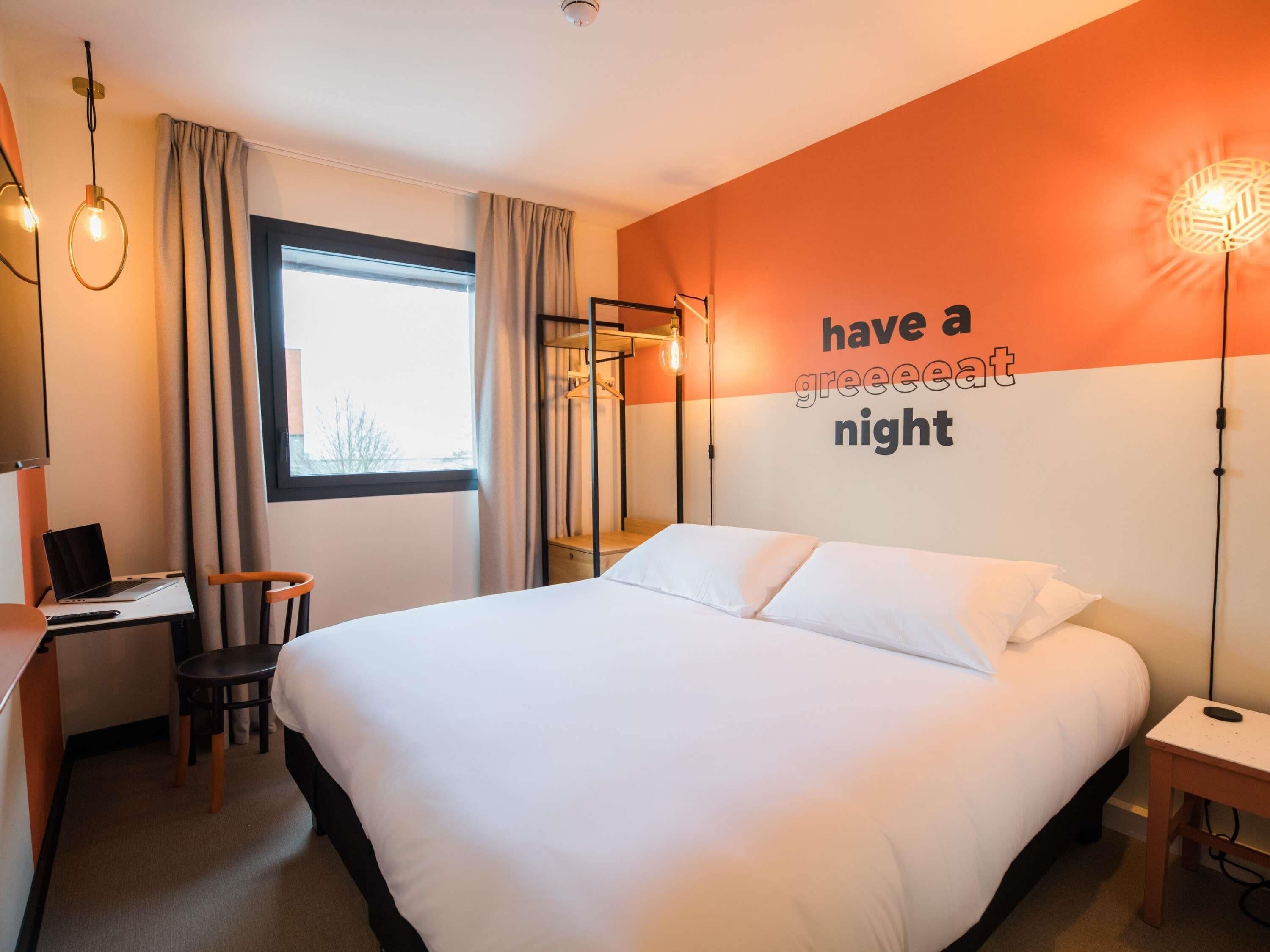Una habitación de hotel con una cama, un escritorio con una computadora portátil, una silla, una mesita de noche y una pared con una cita motivacional.