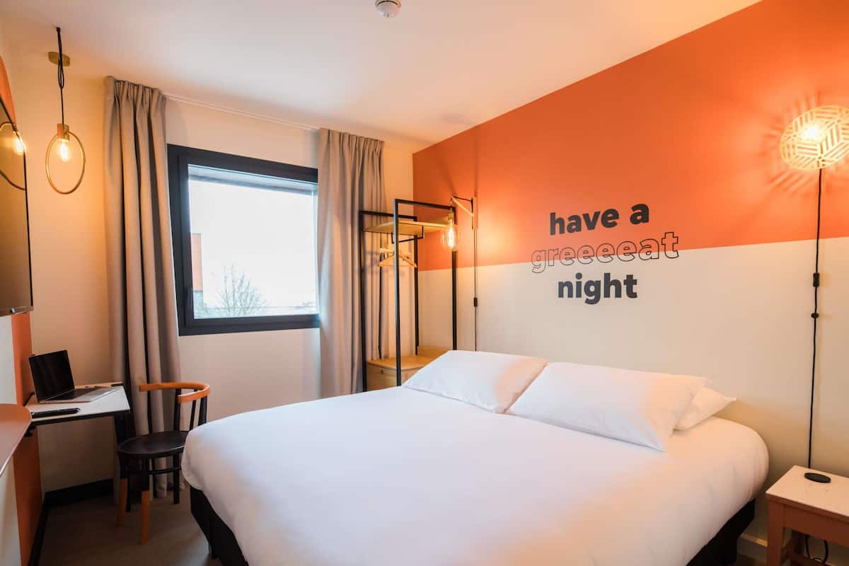 Ein Hotelzimmer mit einem Bett, einem Schreibtisch mit einem Laptop, einem Sessel, einem Nachttisch und einer Wand mit einem motivierenden Zitat.