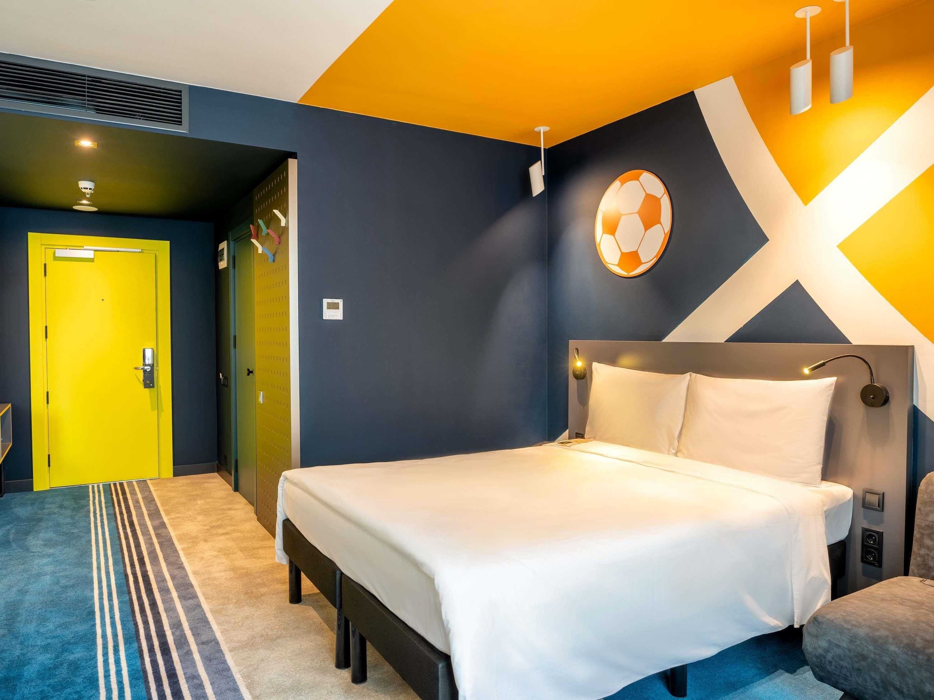 A modern hotel room with a bed, a yellow door, and a unique wall design.
