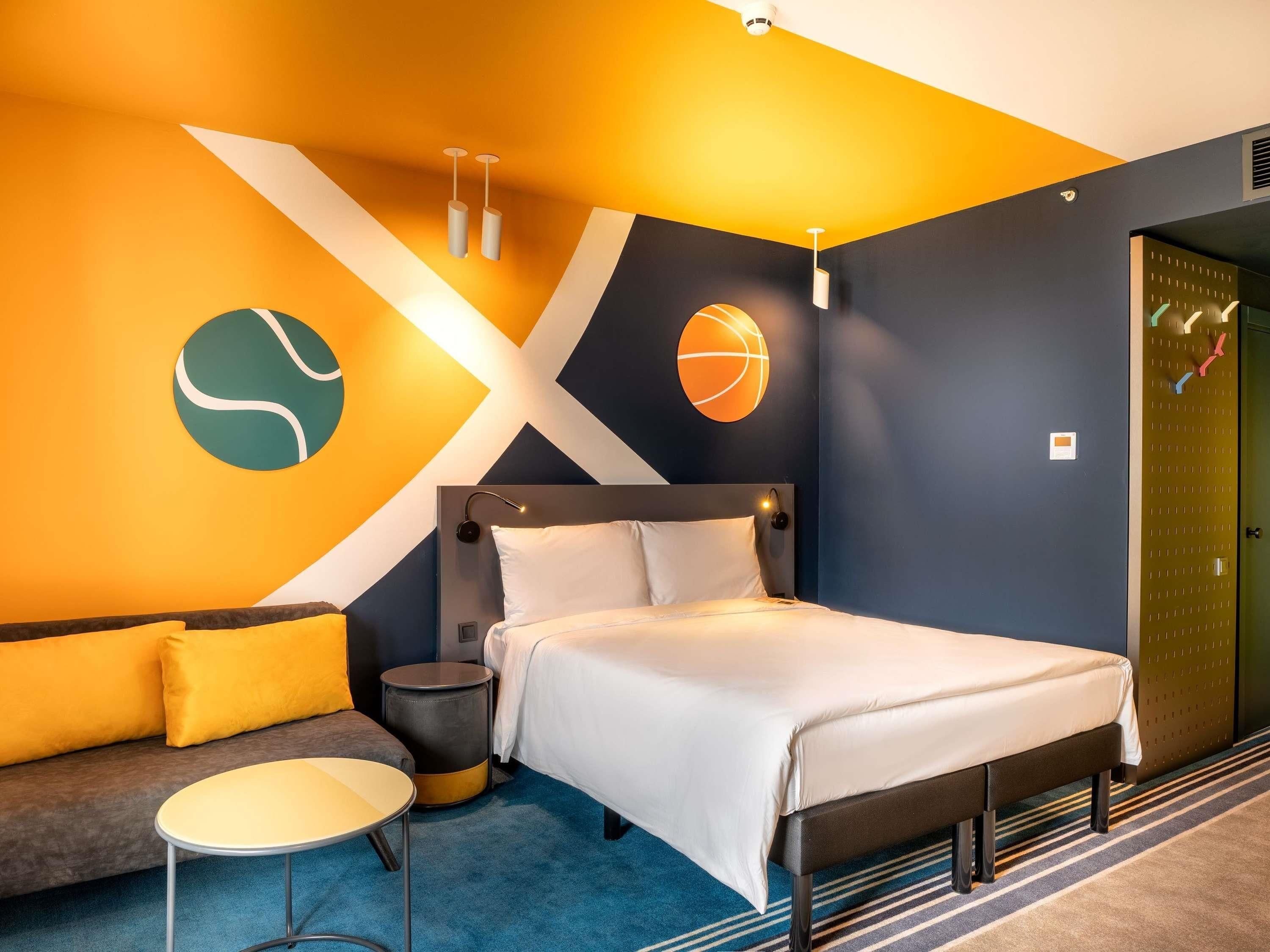 A modern hotel room with a bed, a sofa, a small table, and a wall with geometric patterns.
