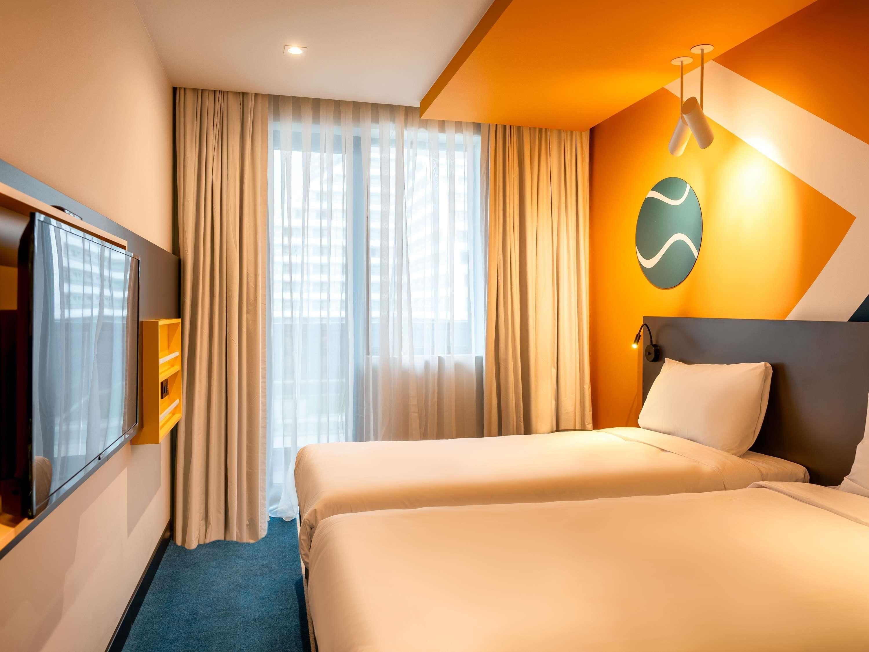 A hotel room with two beds, a TV, a large window with curtains, and a wall-mounted light fixture.