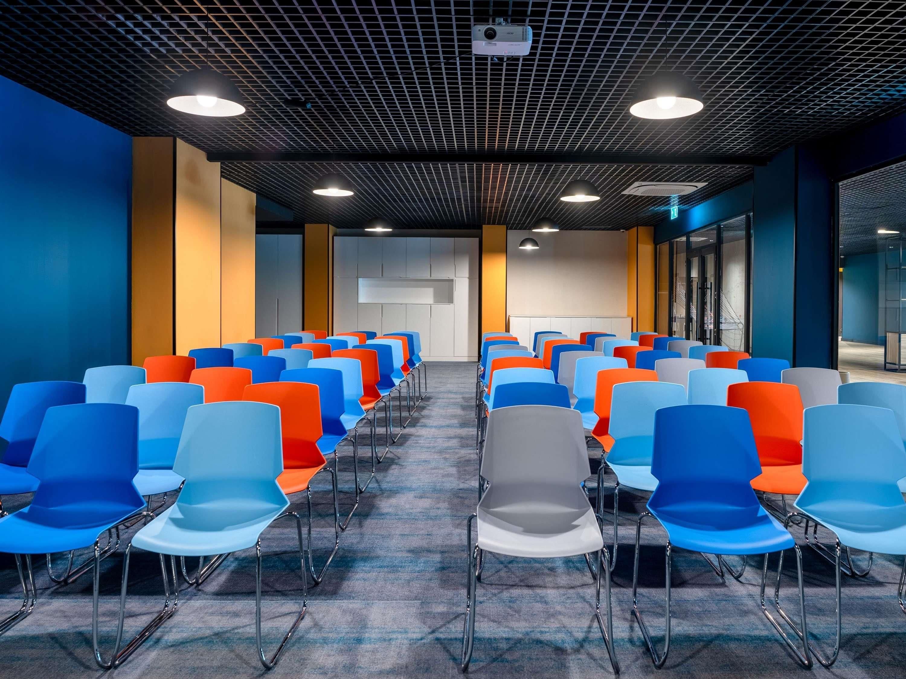 Une salle de conférence avec des rangées de chaises, un projecteur au plafond et une grande fenêtre sur le côté droit.