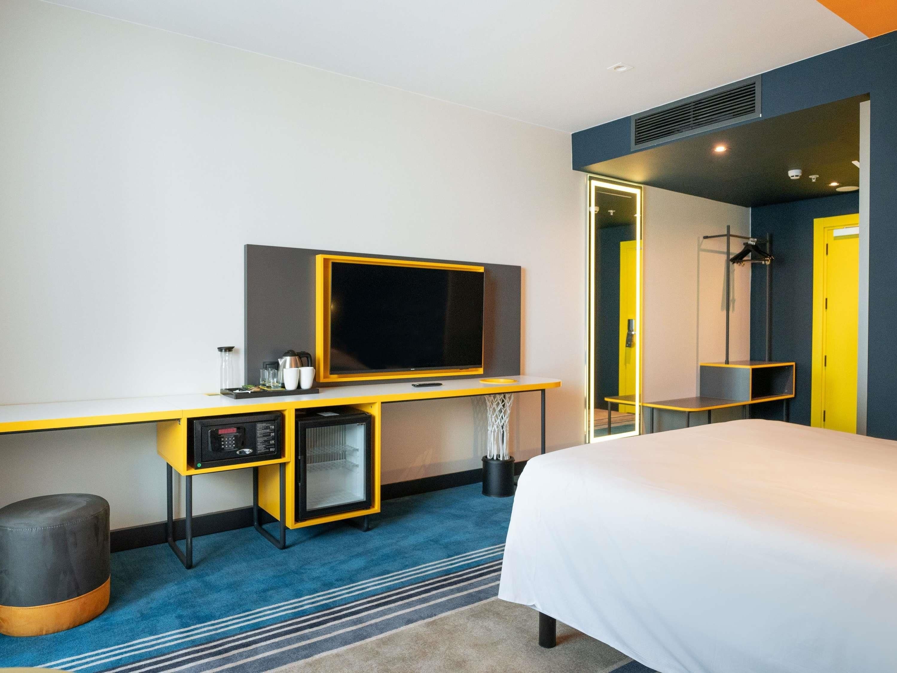 Une chambre d’hôtel équipée d’un lit, d’une télévision, d’une petite kitchenette et d’une salle de bain visible à travers une porte en verre.