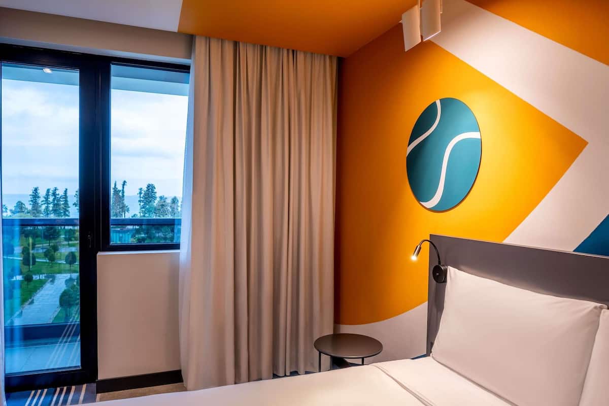 A hotel room with a bed, a small table, a curtain, a window with a view, and a wall with a geometric design.