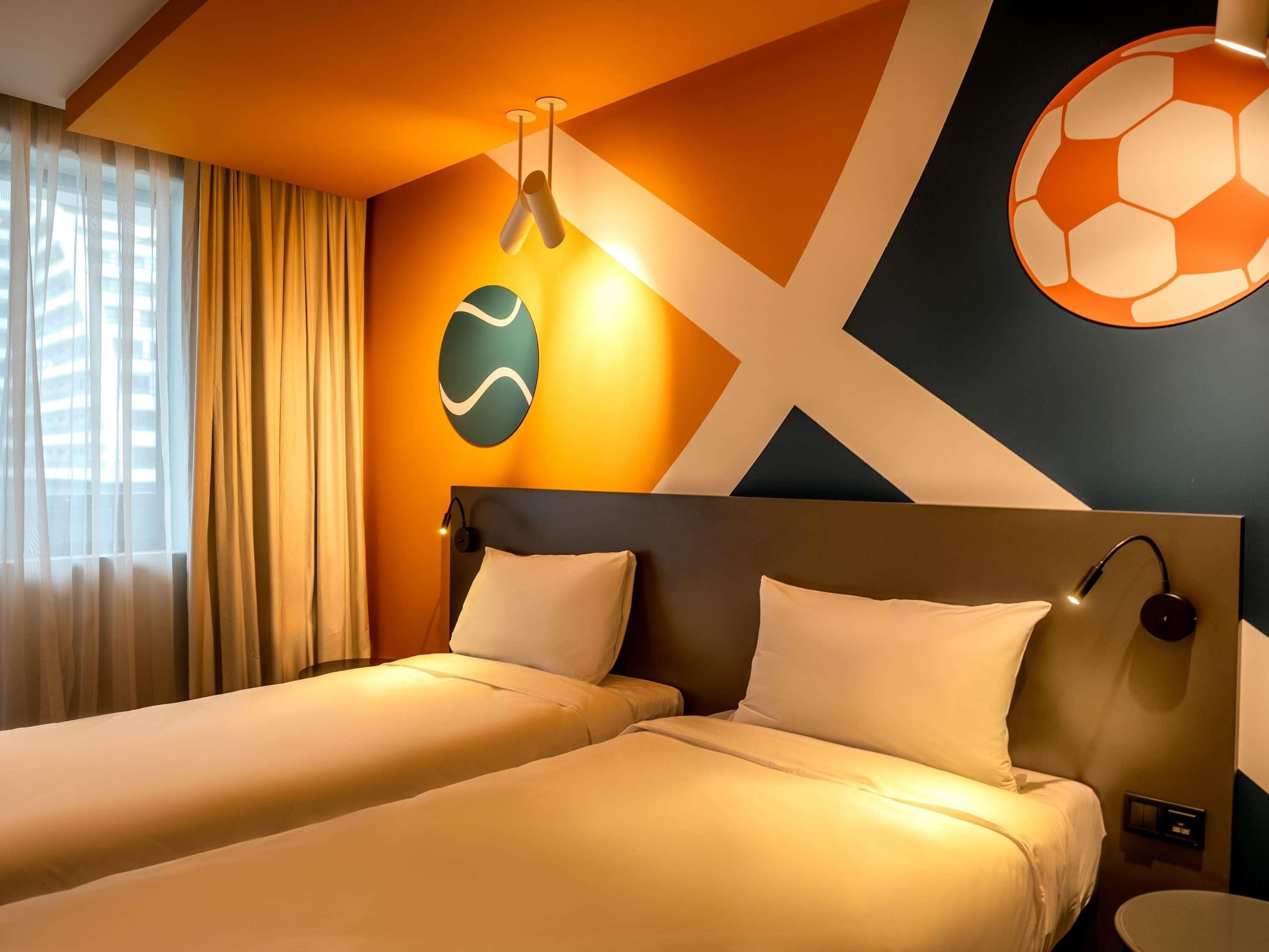 A hotel room with two beds, a large window with curtains, a wall with a geometric pattern, and a wall-mounted light fixture.