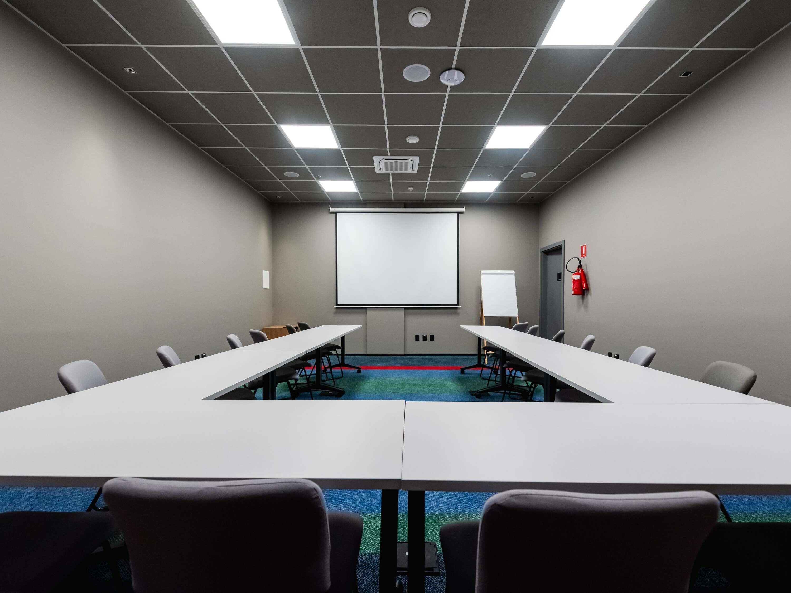 Uma sala de conferência com uma mesa comprida, cadeiras, tela de projetor, quadro branco e extintor de incêndio.