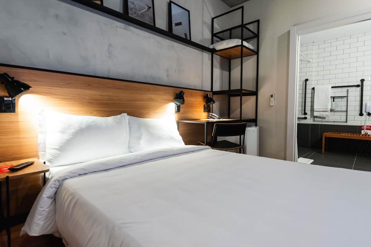 A hotel room with a bed, a desk, a chair, a lamp, a shelf with pictures, and a bathroom with a towel rack.