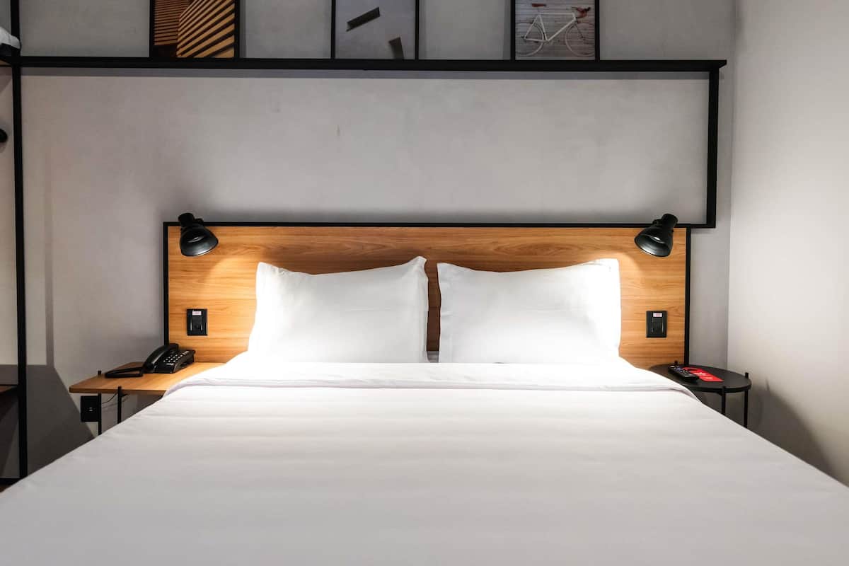 A hotel room with a wooden headboard, white bedding, two bedside lamps, a phone, and a small table with a red item on it.